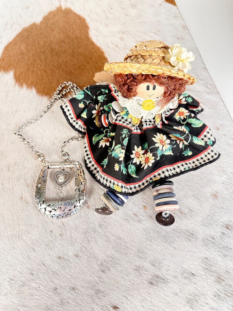 Pode incluir: Uma boneca feita &agrave; m&atilde;o vestindo um vestido floral preto e branco com um chap&eacute;u de palha e uma flor branca. A boneca est&aacute; segurando um colar de corrente de prata com um pingente em forma de cora&ccedil;&atilde;o.