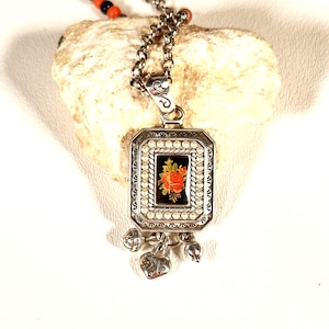 May include: Silver pendant necklace with a rectangular pendant featuring a floral design. The pendant has a black background with a red and orange rose. The necklace has a chain with small beads.