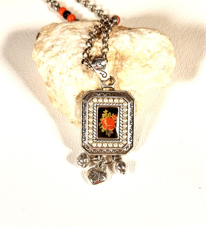 May include: Silver pendant necklace with a rectangular pendant featuring a floral design. The pendant has a black background with a red and orange rose. The necklace has a chain with small beads.
