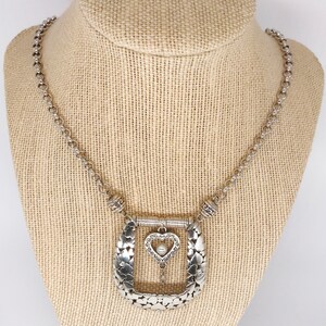 Pode incluir: Um colar de prata com um grande pingente adornado em forma de ferradura. O pingente &eacute; decorado com pequenos cora&ccedil;&otilde;es e um charme de cora&ccedil;&atilde;o pendurado com uma p&eacute;rola no centro.