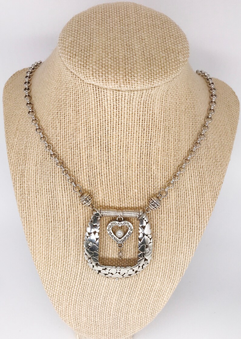 Pode incluir: Um colar de prata com um grande pingente adornado em forma de ferradura. O pingente &eacute; decorado com pequenos cora&ccedil;&otilde;es e um charme de cora&ccedil;&atilde;o pendurado com uma p&eacute;rola no centro.