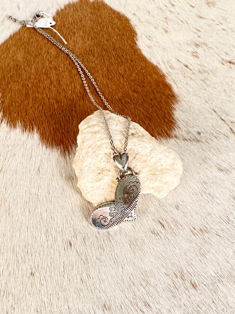 May include: A silver necklace with a heart-shaped pendant. The pendant has a detailed design of swirls and lines.