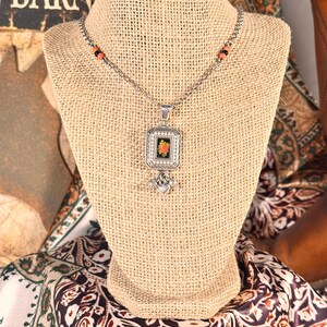 May include: Silver necklace with a square pendant showcasing a floral design. The pendant is set in a decorative frame and hangs from a chain with orange and black beads. The necklace is displayed on a burlap bust.