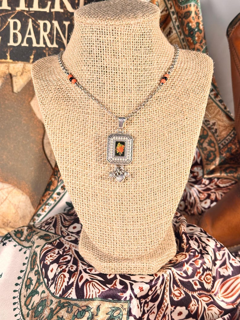 May include: Silver necklace with a square pendant showcasing a floral design. The pendant is set in a decorative frame and hangs from a chain with orange and black beads. The necklace is displayed on a burlap bust.