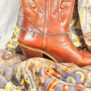 K&ouml;nnte beinhalten: Ein brauner Leder-Cowboystiefel mit dekorativen N&auml;hten und einer silbernen Fu&szlig;kettchen. Der Stiefel liegt auf einem farbenfrohen Paisley-Stoff mit gelben, blauen und orangen Farbt&ouml;nen.
