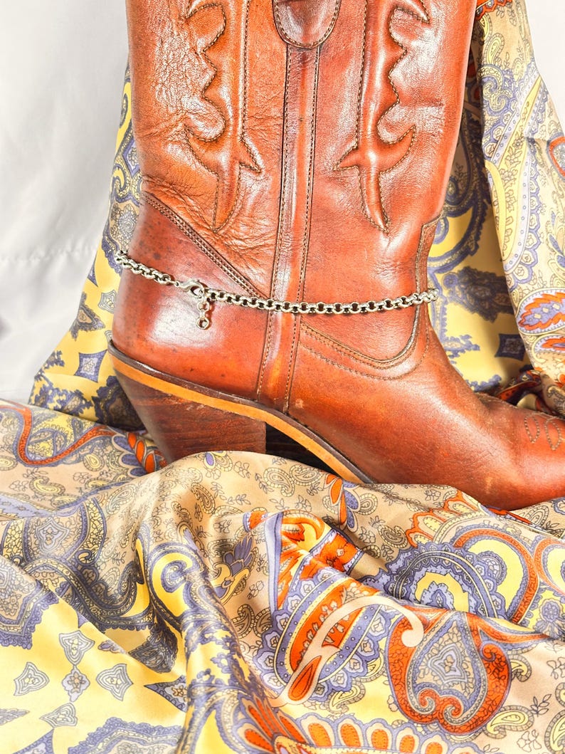 K&ouml;nnte beinhalten: Ein brauner Leder-Cowboystiefel mit dekorativen N&auml;hten und einer silbernen Fu&szlig;kettchen. Der Stiefel liegt auf einem farbenfrohen Paisley-Stoff mit gelben, blauen und orangen Farbt&ouml;nen.
