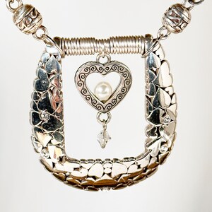 Pode incluir: Colar de prata com um pingente em forma de cora&ccedil;&atilde;o e uma p&eacute;rola. O pingente em forma de cora&ccedil;&atilde;o &eacute; rodeado por uma moldura de prata com um design de espiral. O colar &eacute; feito de uma corrente de pequenos cora&ccedil;&otilde;es de prata.