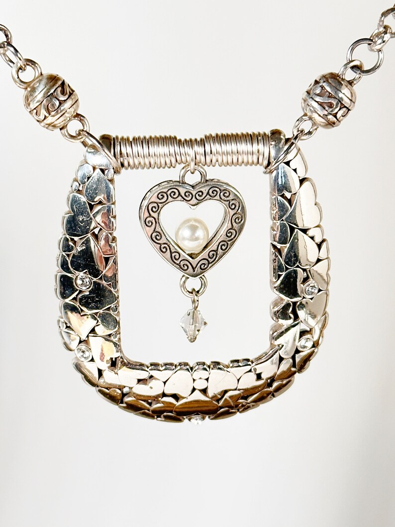 Pode incluir: Colar de prata com um pingente em forma de cora&ccedil;&atilde;o e uma p&eacute;rola. O pingente em forma de cora&ccedil;&atilde;o &eacute; rodeado por uma moldura de prata com um design de espiral. O colar &eacute; feito de uma corrente de pequenos cora&ccedil;&otilde;es de prata.