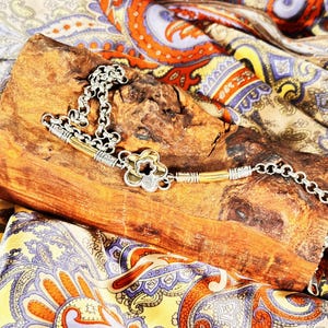 K&ouml;nnte beinhalten: Eine silberfarbene Halskette mit einem bl&uuml;tenf&ouml;rmigen Anh&auml;nger und einem Kettenmuster, die auf einem Holzst&uuml;ck liegt. Die Halskette hat goldfarbene Akzente. Der Hintergrund ist ein farbenfrohes Paisley-Gewebe.