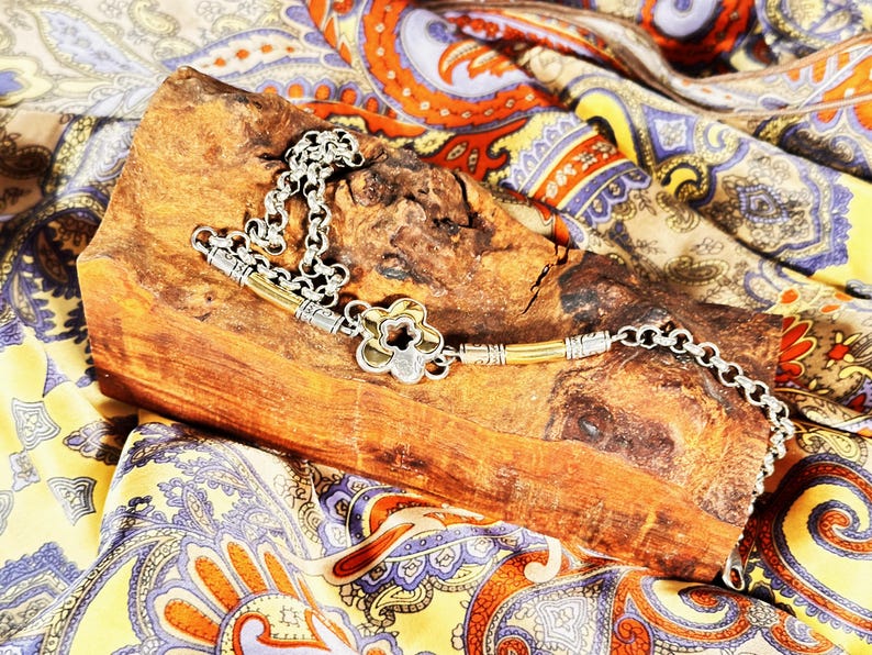 K&ouml;nnte beinhalten: Eine silberfarbene Halskette mit einem bl&uuml;tenf&ouml;rmigen Anh&auml;nger und einem Kettenmuster, die auf einem Holzst&uuml;ck liegt. Die Halskette hat goldfarbene Akzente. Der Hintergrund ist ein farbenfrohes Paisley-Gewebe.