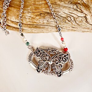 May include: A silver chain necklace with a silver butterfly pendant. The butterfly has intricate details and is surrounded by a silver oval frame. The chain has small silver beads with red, white, and green accents.