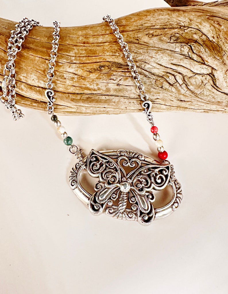 May include: A silver chain necklace with a silver butterfly pendant. The butterfly has intricate details and is surrounded by a silver oval frame. The chain has small silver beads with red, white, and green accents.