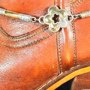 K&ouml;nnte beinhalten: Nahaufnahme eines braunen Lederstiefels mit einer dekorativen silber- und goldfarbenen Stiefelkette. Die Kette hat ein bl&uuml;tenf&ouml;rmiges Mittelst&uuml;ck und aufwendige Details. Der Stiefel hat sichtbare N&auml;hte.