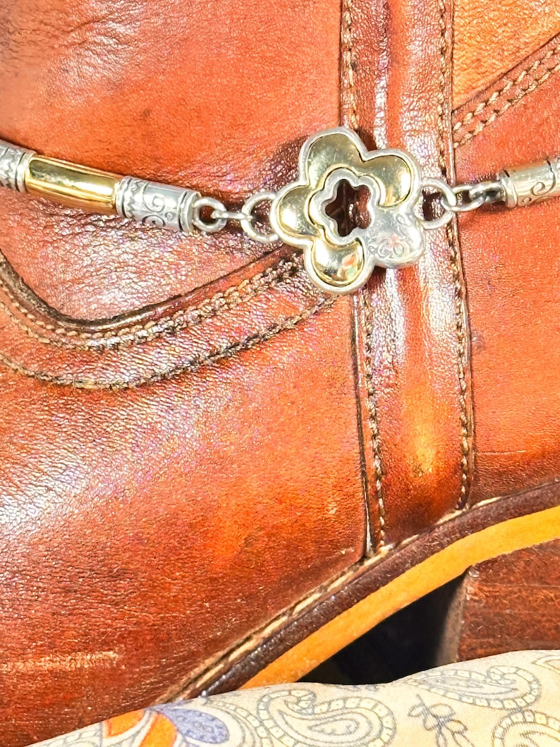 K&ouml;nnte beinhalten: Nahaufnahme eines braunen Lederstiefels mit einer dekorativen silber- und goldfarbenen Stiefelkette. Die Kette hat ein bl&uuml;tenf&ouml;rmiges Mittelst&uuml;ck und aufwendige Details. Der Stiefel hat sichtbare N&auml;hte.