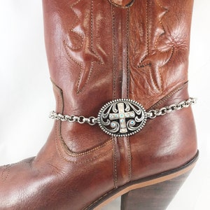 May include: A brown leather cowboy boot with a silver chain and a silver charm with a cross and rhinestones attached to the top of the boot.
