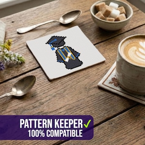 May include: A square, white coaster with a cross-stitch design of a graduate in a cap and gown. The coaster is on a wooden table, next to a cup of coffee, sugar cubes, and flowers. The text "PATTERN KEEPER 100% COMPATIBLE" is at the bottom.