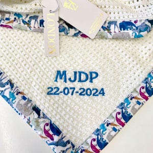 May include: White knitted baby blanket with a blue and white animal print border. The blanket is personalized with the name "MJDP" and the date "22-07-2024" in blue embroidery.