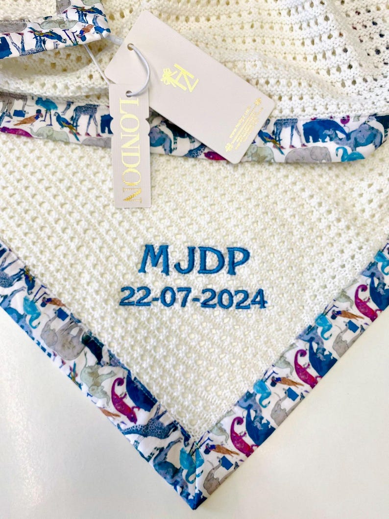May include: White knitted baby blanket with a blue and white animal print border. The blanket is personalized with the name "MJDP" and the date "22-07-2024" in blue embroidery.
