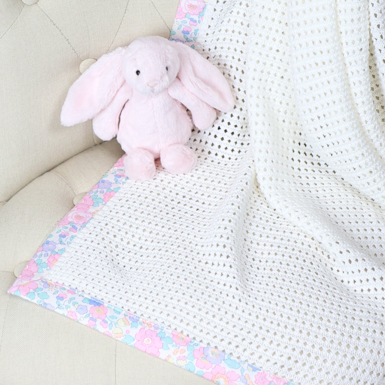 May include: A white crocheted baby blanket with a floral print border. A pink plush bunny is sitting on a white chair in the background.