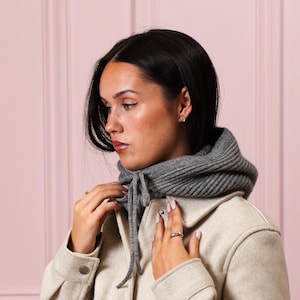 May include: A gray knit hood with a drawstring closure is worn over a light-colored jacket. The hood is ribbed and appears to be made of a soft, warm material. The jacket has large buttons and a classic design.