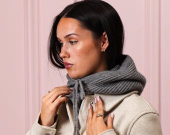 May include: A gray knit hood with a drawstring closure is worn over a light-colored jacket. The hood is ribbed and appears to be made of a soft, warm material. The jacket has large buttons and a classic design.