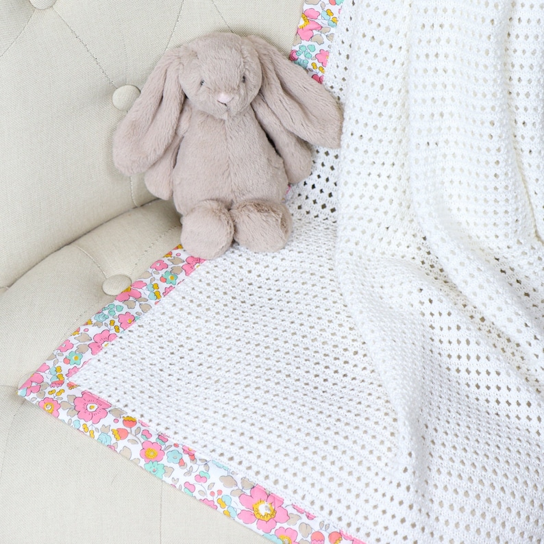 May include: A white crocheted baby blanket with a floral trim. The blanket is folded and placed on a light brown chair with a stuffed bunny rabbit on top.