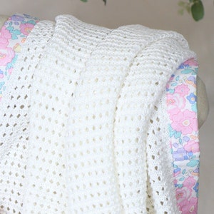 May include: White crocheted baby blanket with a floral print border. The blanket is made with a delicate open stitch pattern.