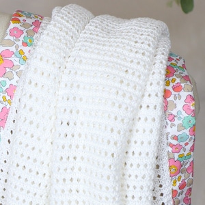 May include: A white crocheted baby blanket with a floral print border. The blanket is made with a delicate open stitch pattern.