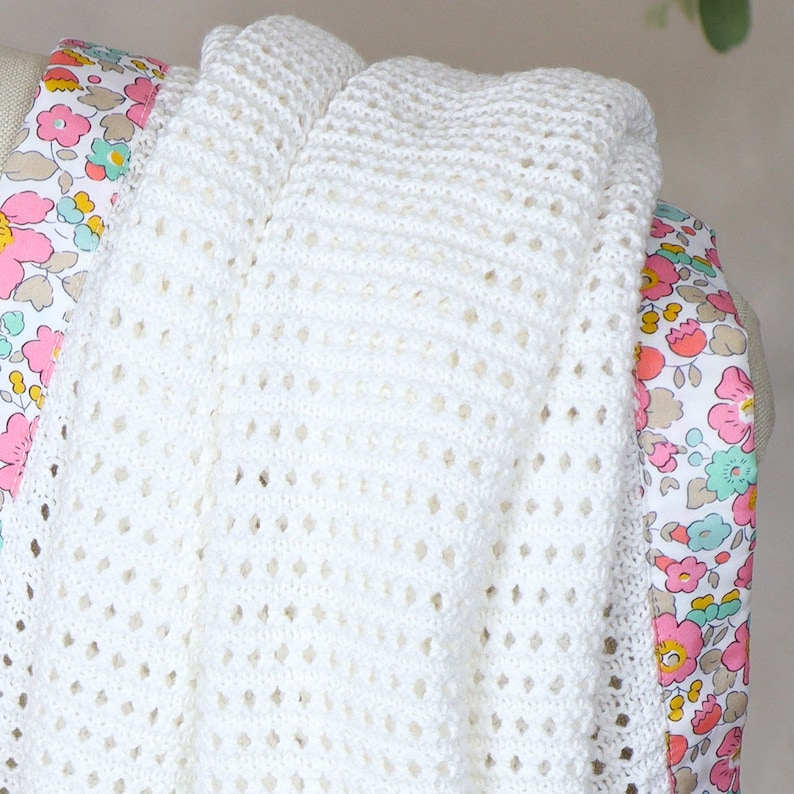 May include: A white crocheted baby blanket with a floral print border. The blanket is made with a delicate open stitch pattern.
