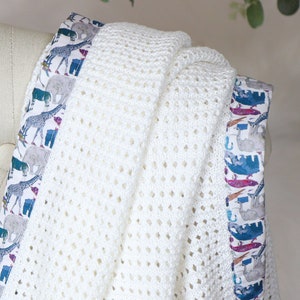 May include: White crochet baby blanket with a colorful animal print border. The border features giraffes, elephants, and other animals.