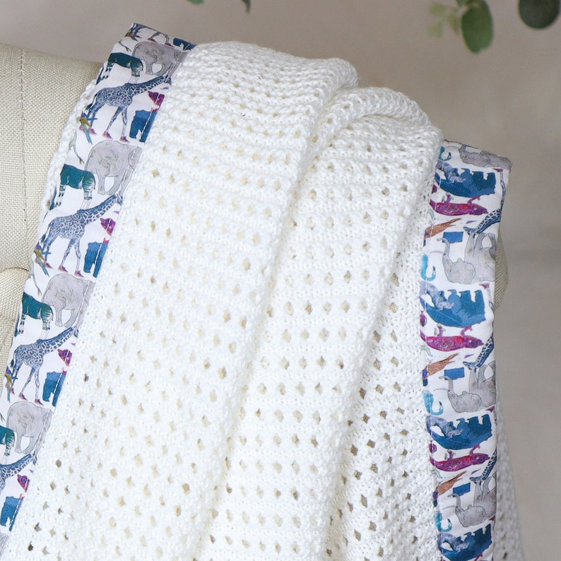 May include: White crochet baby blanket with a colorful animal print border. The border features giraffes, elephants, and other animals.