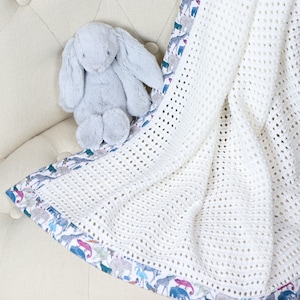 May include: A white crocheted baby blanket with a colorful animal print border. A gray stuffed bunny is sitting on top of the blanket.