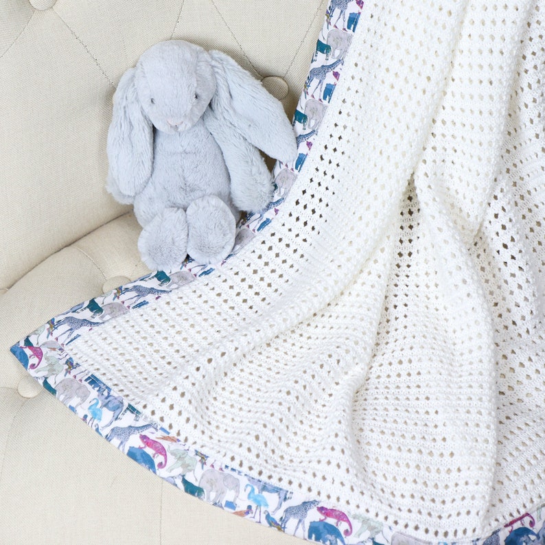 May include: A white crocheted baby blanket with a colorful animal print border. A gray stuffed bunny is sitting on top of the blanket.