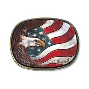 May include: Oval metal belt buckle featuring an American flag design with an eagle and the text "Sitwell". The flag has red and white stripes with a blue field and white stars. The eagle is brown and white.