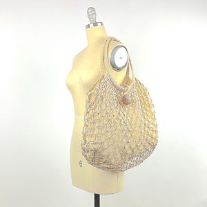 May include: A large, beige, crocheted mesh tote bag with a wooden button closure. The bag is hanging on a mannequin.