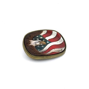 May include: Oval metal belt buckle featuring a painted American flag and a bald eagle. The buckle has a gold-coloured frame. The flag design includes red and white stripes with a blue field of stars. The eagle is brown and white.