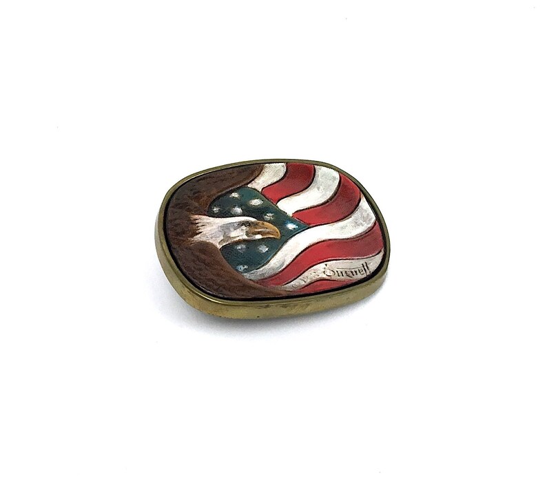 May include: Oval metal belt buckle featuring a painted American flag and a bald eagle. The buckle has a gold-coloured frame. The flag design includes red and white stripes with a blue field of stars. The eagle is brown and white.