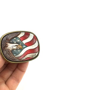 May include: Rectangular belt buckle with a patriotic design. The buckle features a detailed relief of an American flag and a bald eagle. The flag has red and white stripes with a blue field of stars. The eagle is depicted with a detailed beak and feathers.