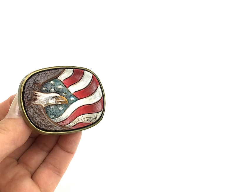 May include: Rectangular belt buckle with a patriotic design. The buckle features a detailed relief of an American flag and a bald eagle. The flag has red and white stripes with a blue field of stars. The eagle is depicted with a detailed beak and feathers.