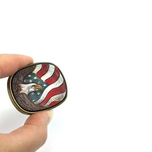 May include: A vintage, square belt buckle with a patriotic design. The buckle features a detailed depiction of an American bald eagle and the U.S. flag in red, white, and blue. The buckle has a bronze-coloured frame.