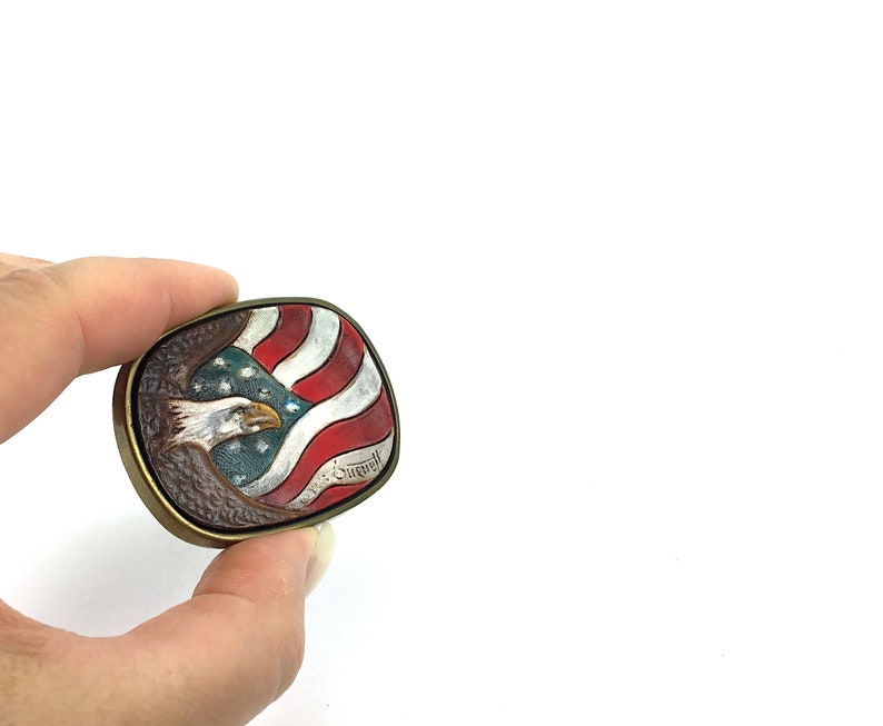 May include: A vintage, square belt buckle with a patriotic design. The buckle features a detailed depiction of an American bald eagle and the U.S. flag in red, white, and blue. The buckle has a bronze-coloured frame.