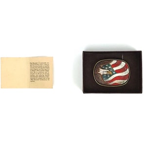 May include: Oval belt buckle featuring an American flag design with a bald eagle. The buckle is set in a brown leather frame. The eagle is painted with blue, white, and red colours. The buckle is in a brown box.