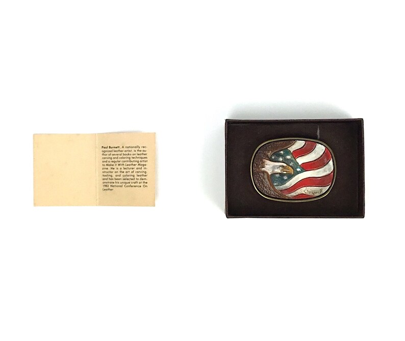 May include: Oval belt buckle featuring an American flag design with a bald eagle. The buckle is set in a brown leather frame. The eagle is painted with blue, white, and red colours. The buckle is in a brown box.