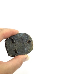 May include: A vintage brass belt buckle with an oval shape. The buckle has a tarnished, aged appearance with visible text and a pin for attachment. The back of the buckle shows signs of wear and use.