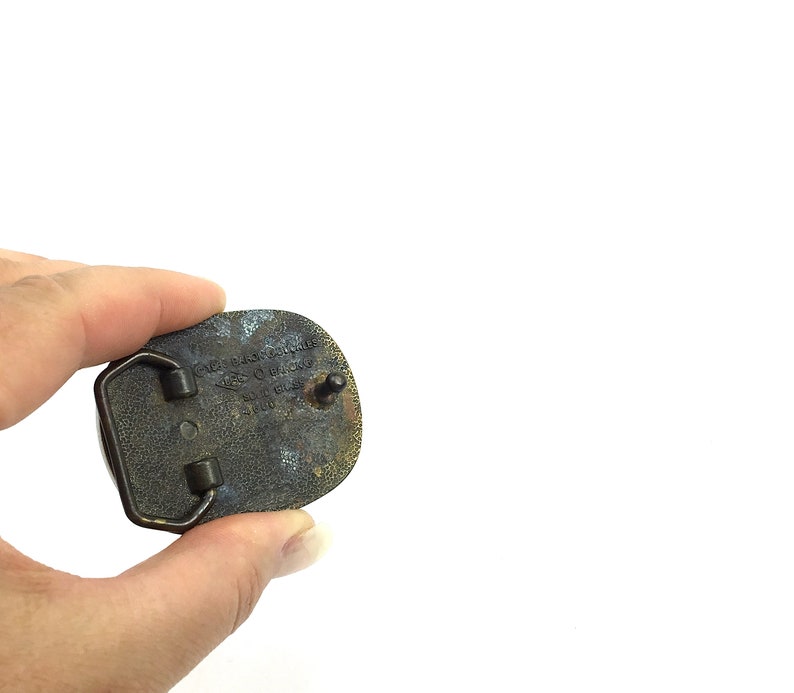 May include: A vintage brass belt buckle with an oval shape. The buckle has a tarnished, aged appearance with visible text and a pin for attachment. The back of the buckle shows signs of wear and use.
