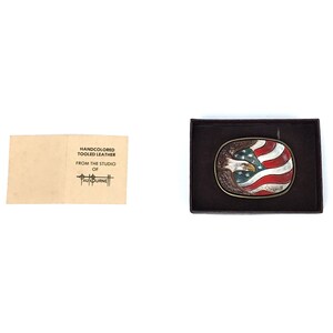 May include: Oval belt buckle featuring an American flag design with a bald eagle. The buckle is in a dark brown box. The flag has red and white stripes with a blue field and white stars. The eagle is brown and white. A small card reads "HANDCOLORED TOOLED LEATHER FROM THE STUDIO OF PAUL BURNE".