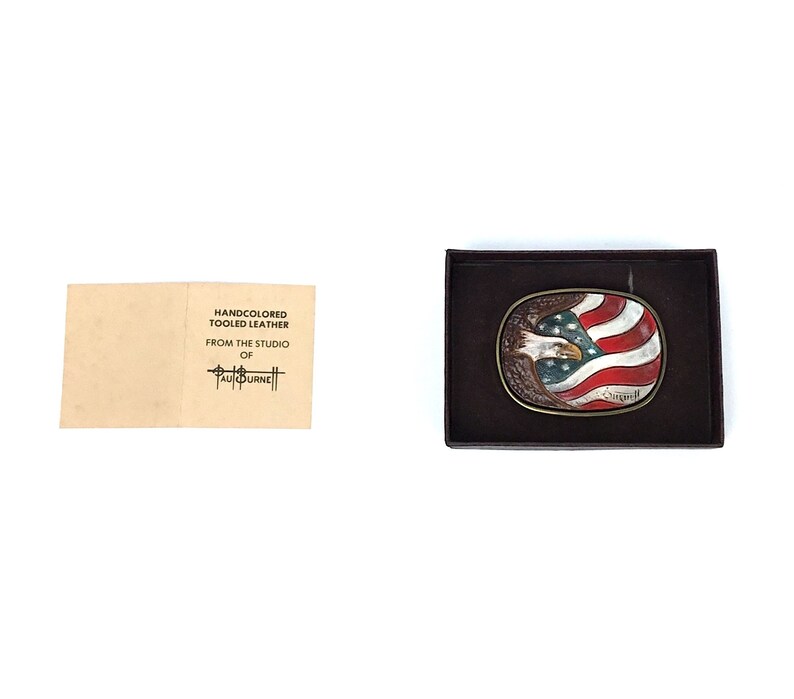 May include: Oval belt buckle featuring an American flag design with a bald eagle. The buckle is in a dark brown box. The flag has red and white stripes with a blue field and white stars. The eagle is brown and white. A small card reads "HANDCOLORED TOOLED LEATHER FROM THE STUDIO OF PAUL BURNE".