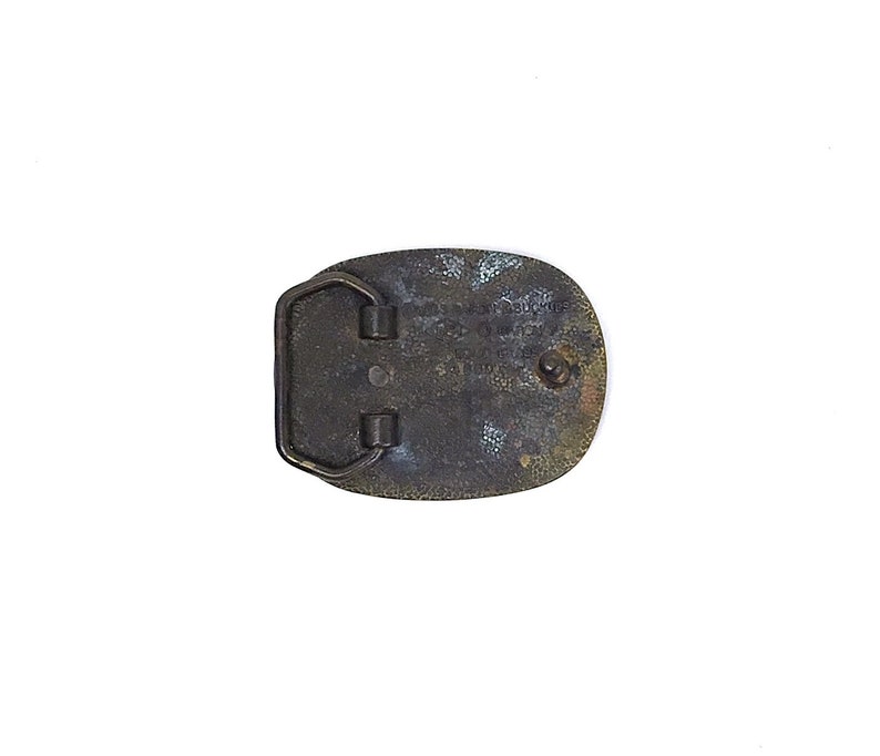 May include: Vintage oval belt buckle with a dark bronze finish. The buckle has a rectangular bar and two loops for the belt. The back of the buckle has engraved text. The buckle is likely made of metal.