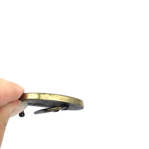 May include: A vintage brass belt buckle with a circular design. The buckle has a dark, textured centre and a lighter, polished brass rim. The buckle's pin and loop are visible.