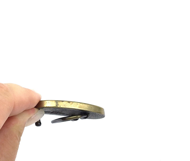 May include: A vintage brass belt buckle with a circular design. The buckle has a dark, textured centre and a lighter, polished brass rim. The buckle's pin and loop are visible.